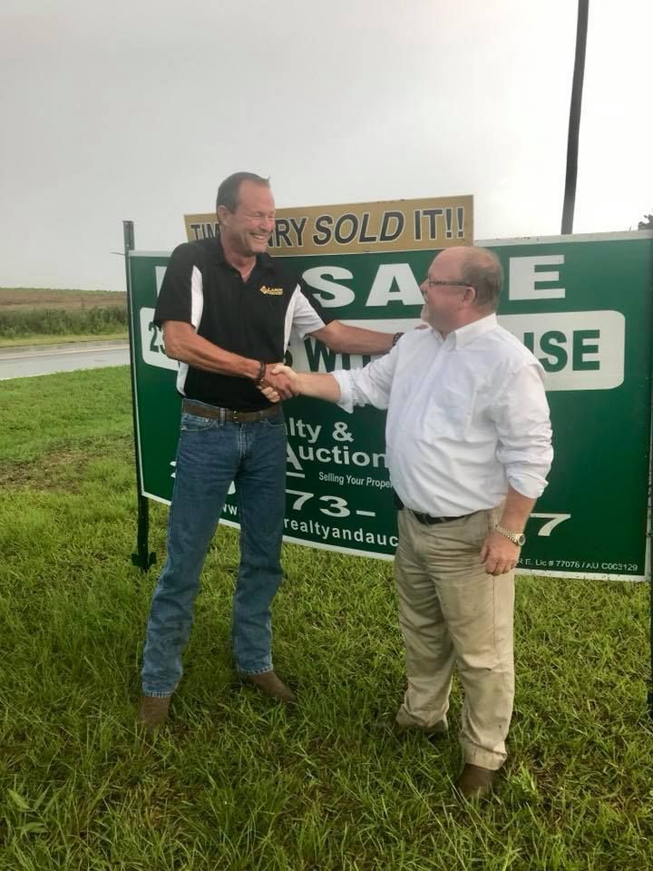 Two people in casual attire shake hands in front of a green real estate sign that says 