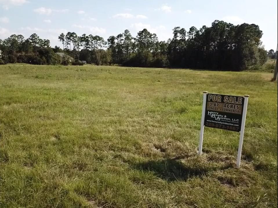A grassy vacant lot with a 'For Sale' sign on the right, backed by a line of trees under a clear, bright sky.