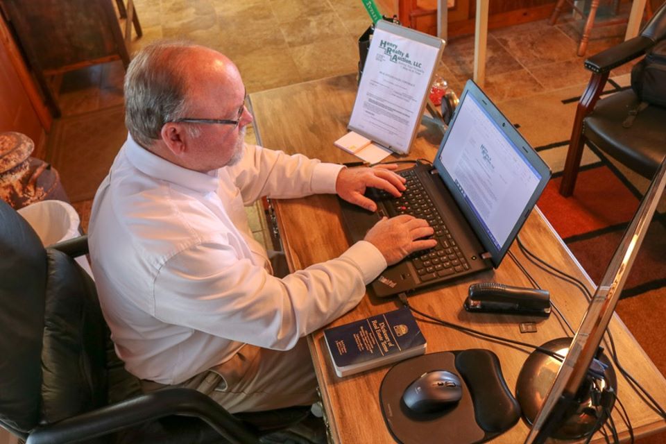 A person in a white button-down shirt works on a laptop at a wooden desk with a mouse, mousepad, and a booklet.