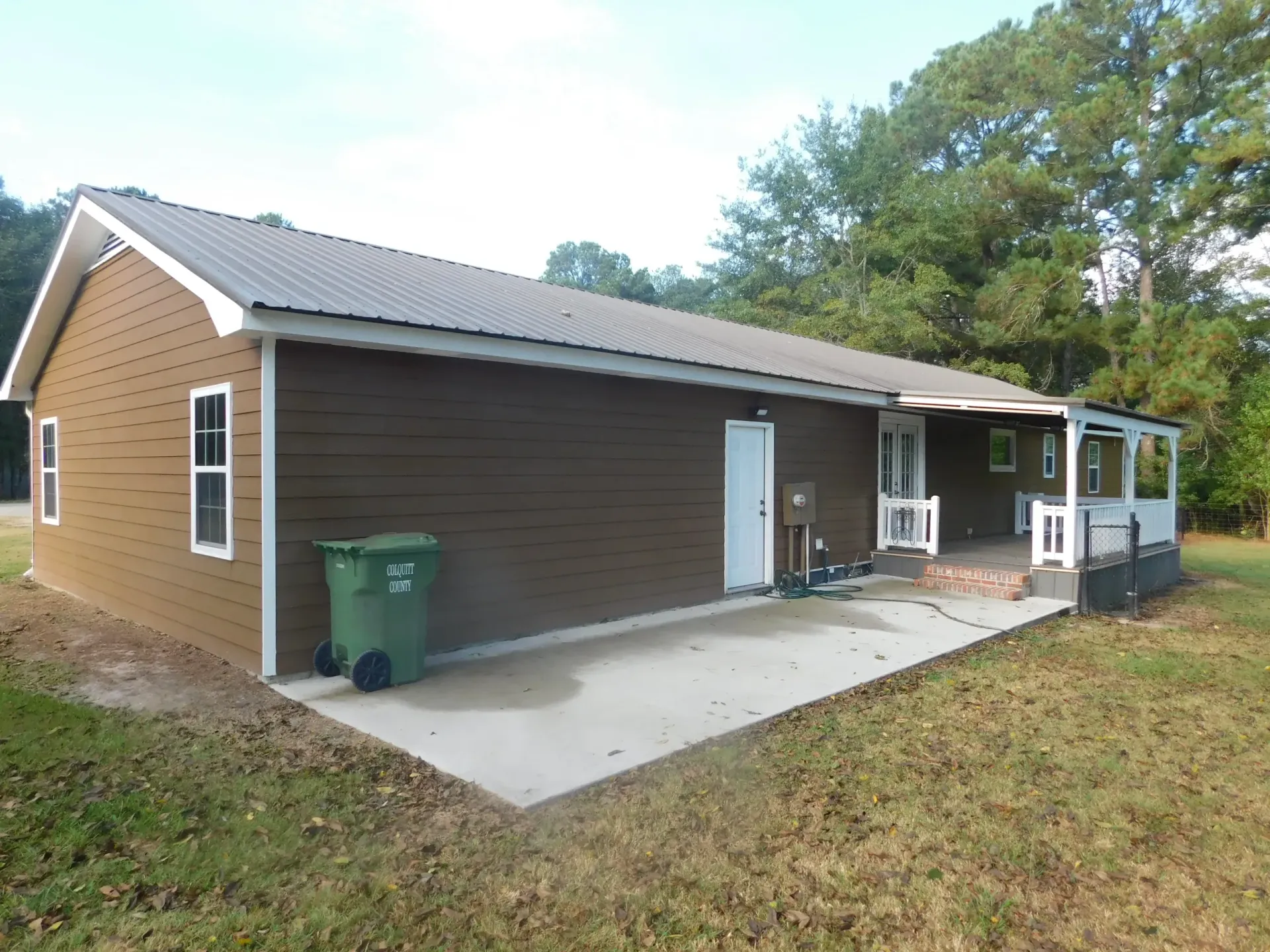 A side view of a brown, one-story house with a metal roof, a white door, and a small covered porch in a grassy yard.