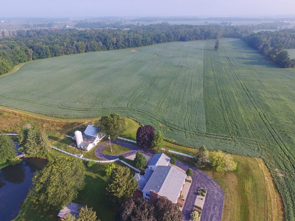 An aerial view of a farm property featuring a house, a silo, a pond, and expansive green fields bordered by woods.