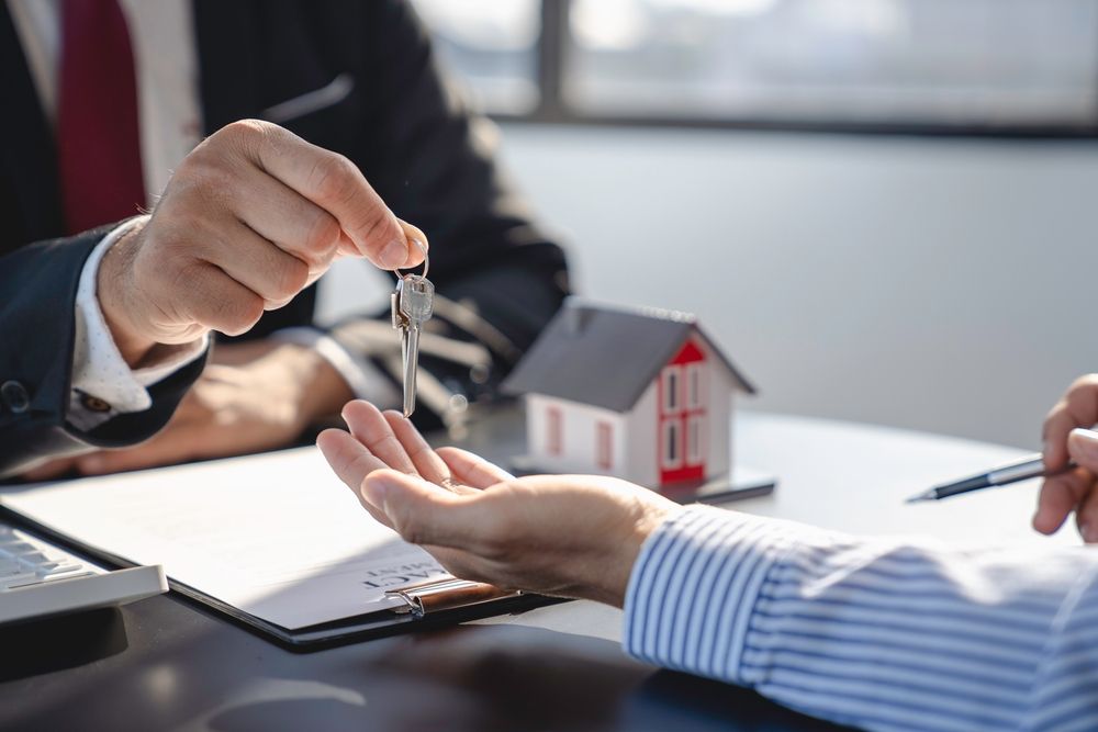 A professional handing a set of house keys to another person across a desk with a small model house and documents.