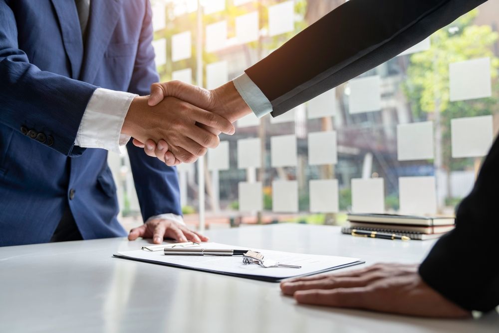 Two business professionals in suits shake hands over a desk with documents, symbolizing a finalized agreement.
