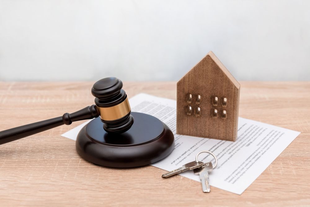 A wooden gavel, a miniature house model, and keys resting on a legal document on a wooden table.