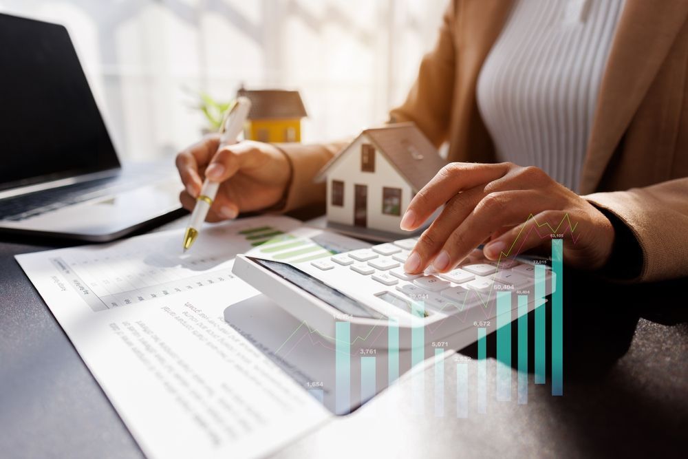 A person calculates real estate finances with a calculator, documents, and a miniature house model on a desk.
