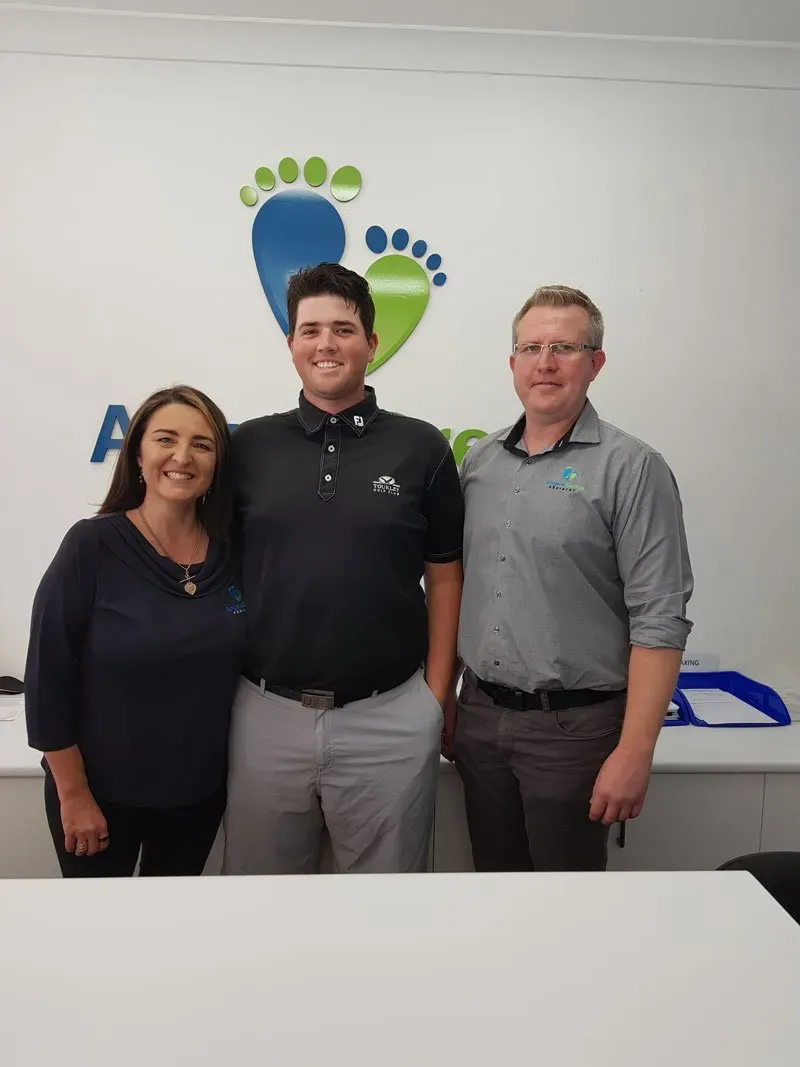 Three people smiling and standing together in an office in front of a wall logo.