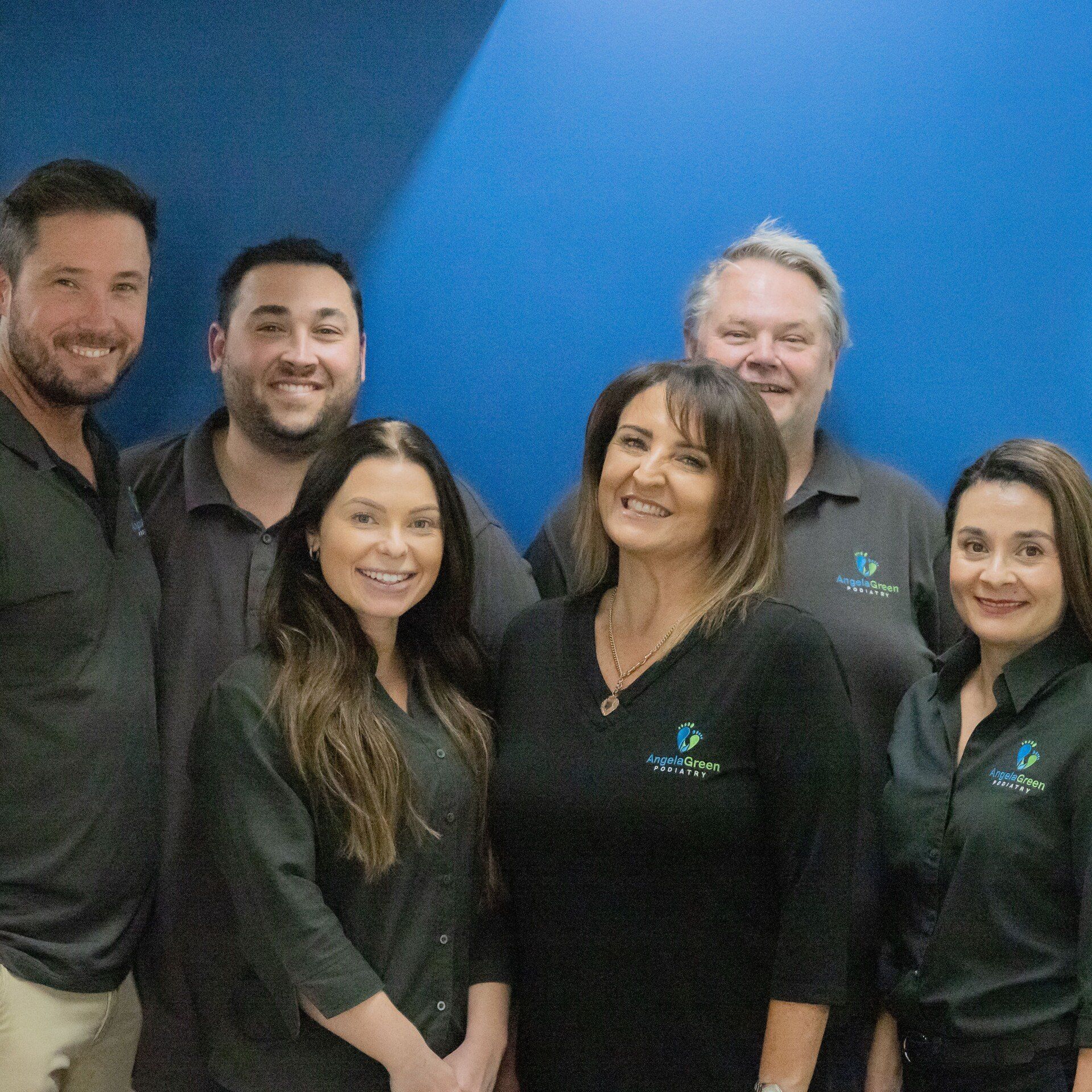 Six coworkers posing in matching black polos against a blue wall, smiling at the camera