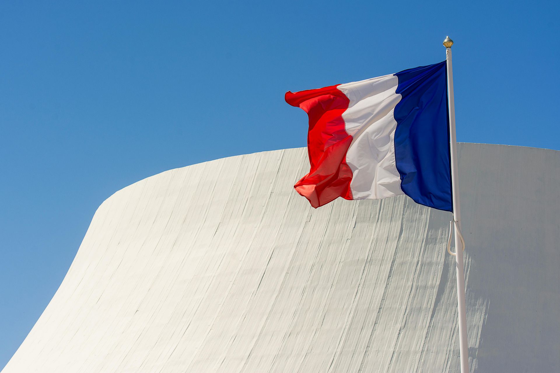 Drapeau français flottant sur un ciel bleu éclatant devant un bâtiment blanc.