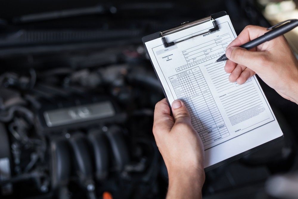 A Person Is Writing On A Clipboard In Front Of A Car Engine — Bay City Quality Mobile Mechanic In Pialba, QLD
