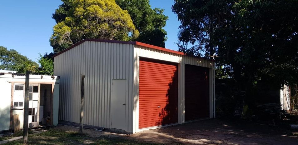 A White Garage With A Red Door Is Surrounded By Trees — Bushman Sheds in Bundaberg East, QLD