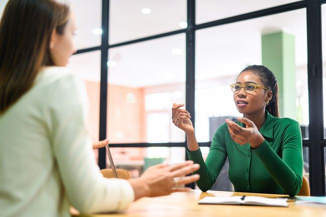 Two people discussing across a glass table in a bright office, one gesturing while speaking.