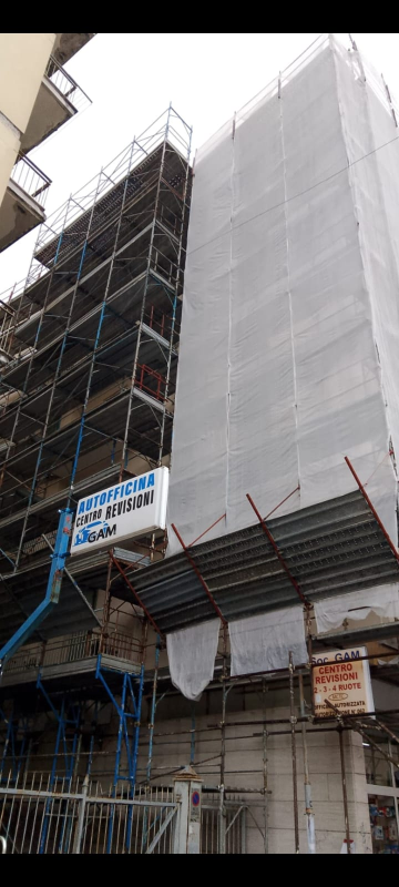 Impalcatura su un edificio avvolta in rete di sicurezza bianca, con supporti da costruzione blu sottostanti.