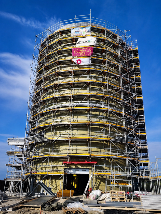 Edificio cilindrico in costruzione, con impalcature, sullo sfondo di un cielo azzurro.