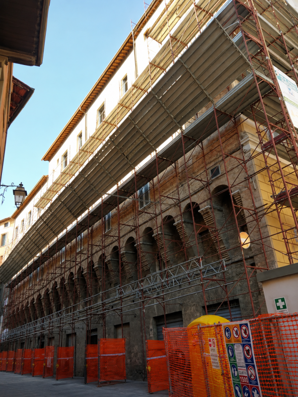 Facciata di un edificio con impalcature su una strada cittadina, con barriere di sicurezza arancioni e un cielo azzurro limpido.