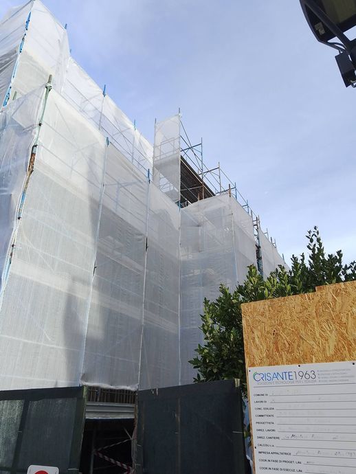 Impalcature argentate da cantiere attorno a un edificio in ristrutturazione, accanto a un albero e a una recinzione.