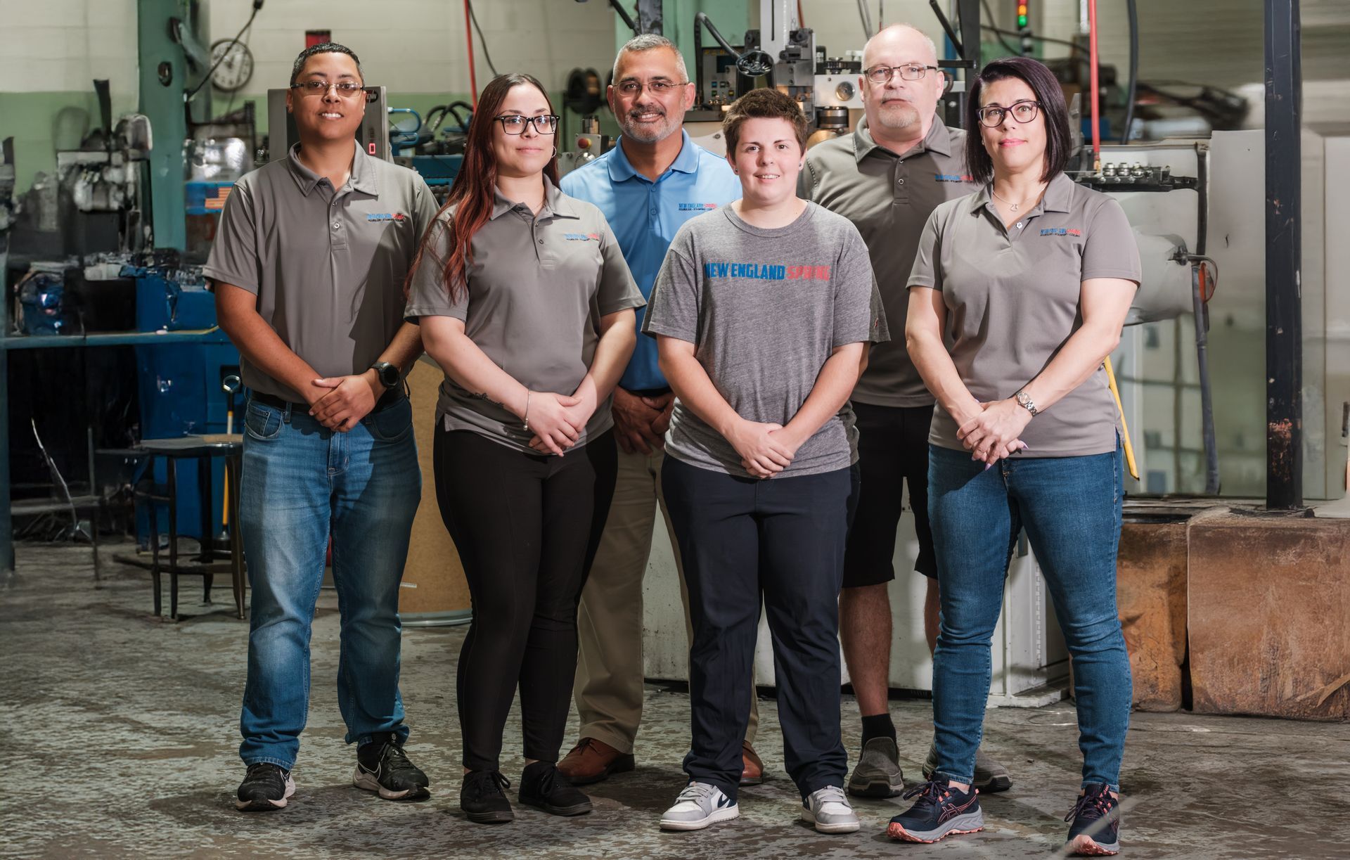 A group of six New England Spring enmployees in gray shirts pose in a workshop.