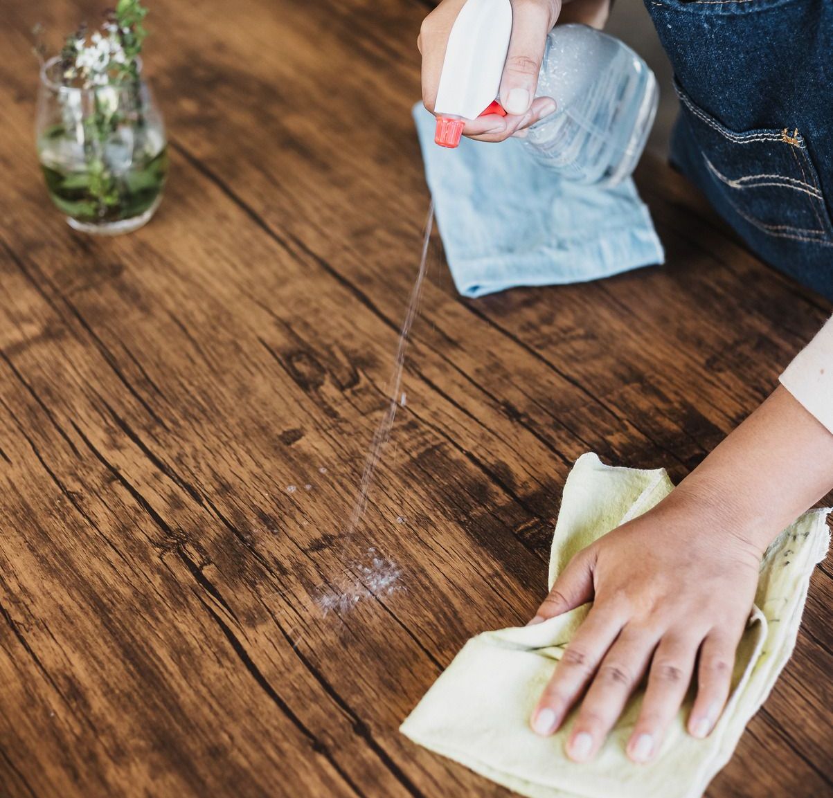 A person is cleaning a wooden table with a cloth and spray bottle