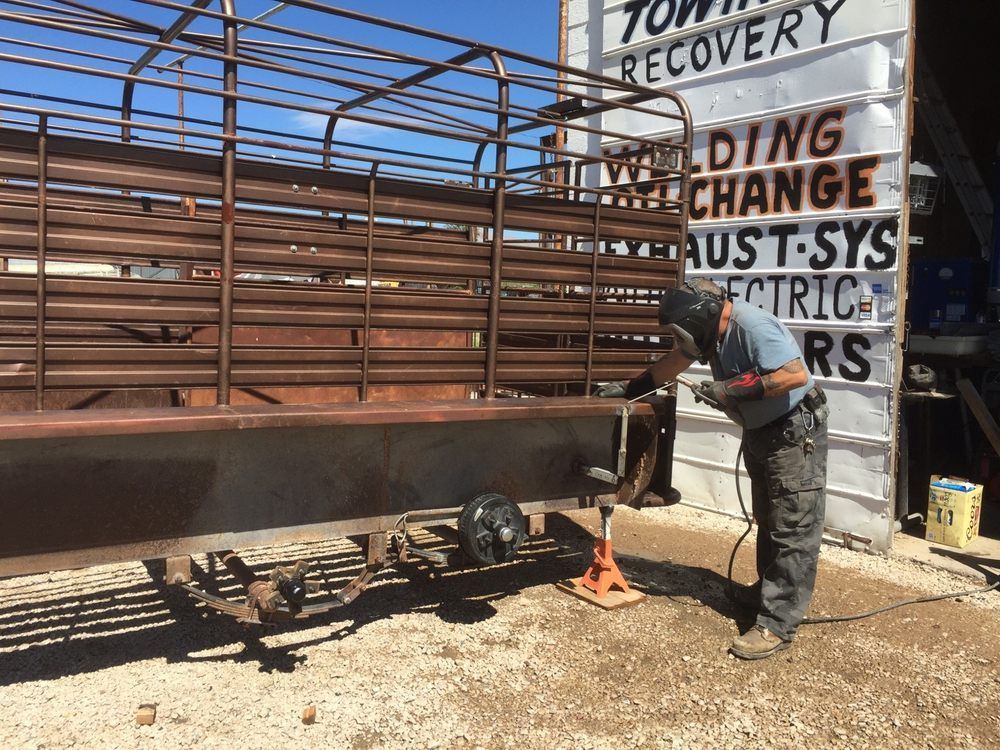 Man welds on trailer frame, outside a repair shop. Trailer is brown, shop sign has text. Sunny day.