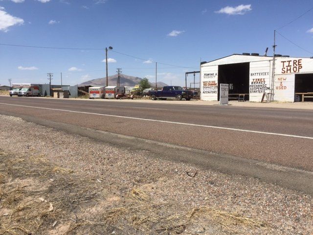 Roadside tire shop, white building with open bay. Gray asphalt road, blue sky, desert landscape.