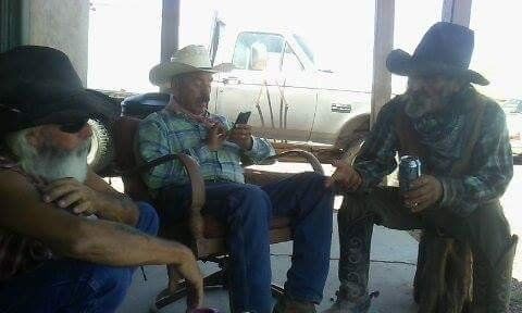 Three cowboys sitting outside. One looks at a phone, one holds a can, one has a long beard and sunglasses.