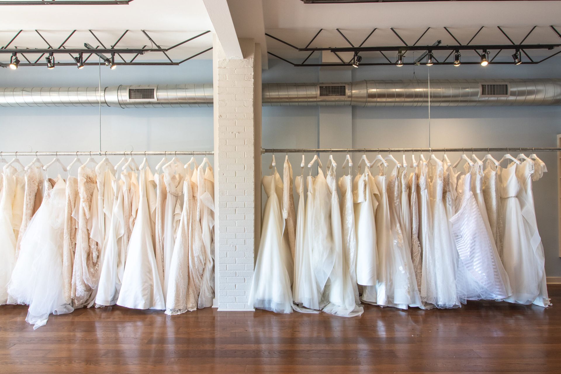 Two rows of white wedding dresses hanging on racks in a bright store with wood floors and industrial lighting.