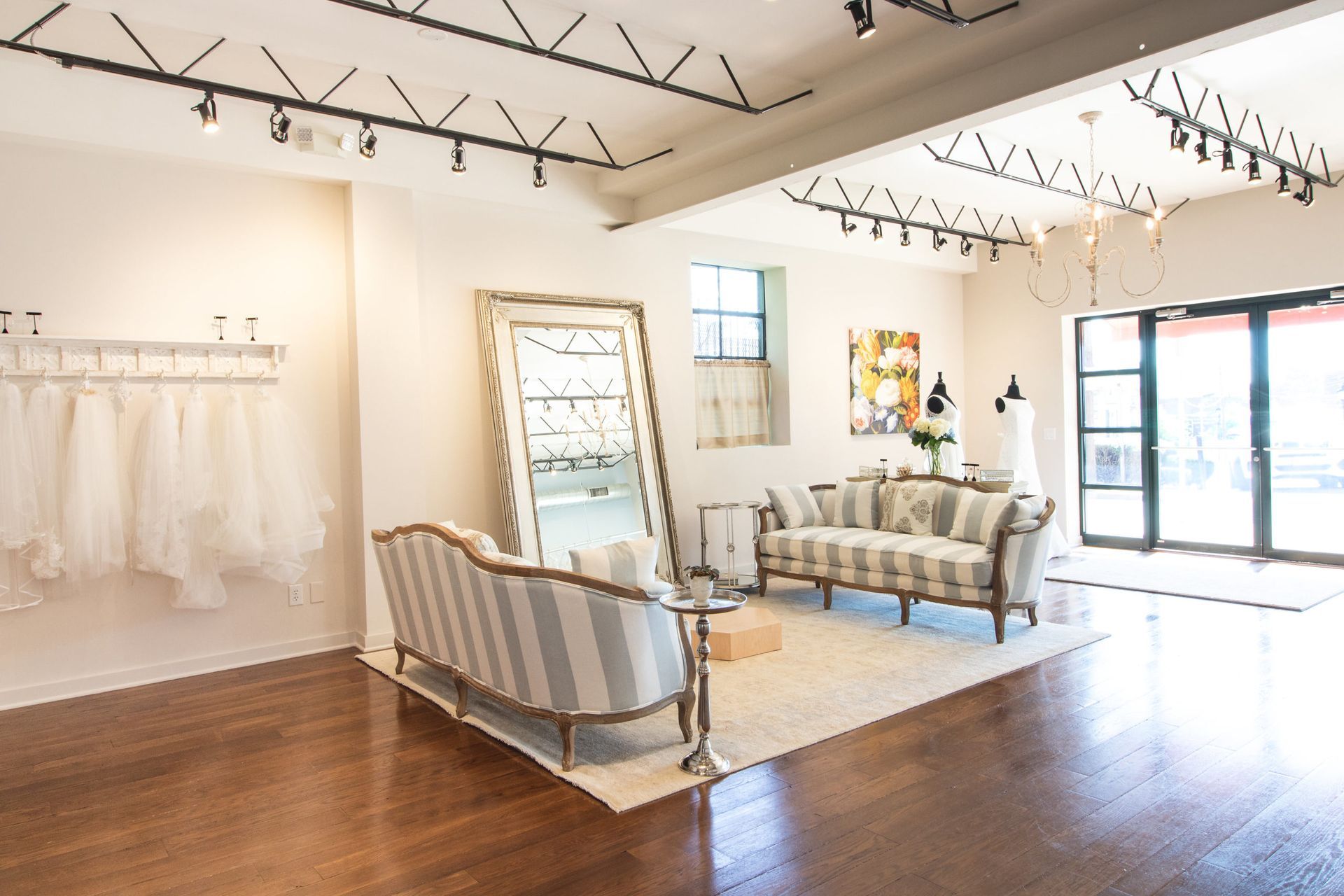 A bright bridal boutique interior with striped sofas, a large ornate mirror, hanging wedding gowns, and track lighting.