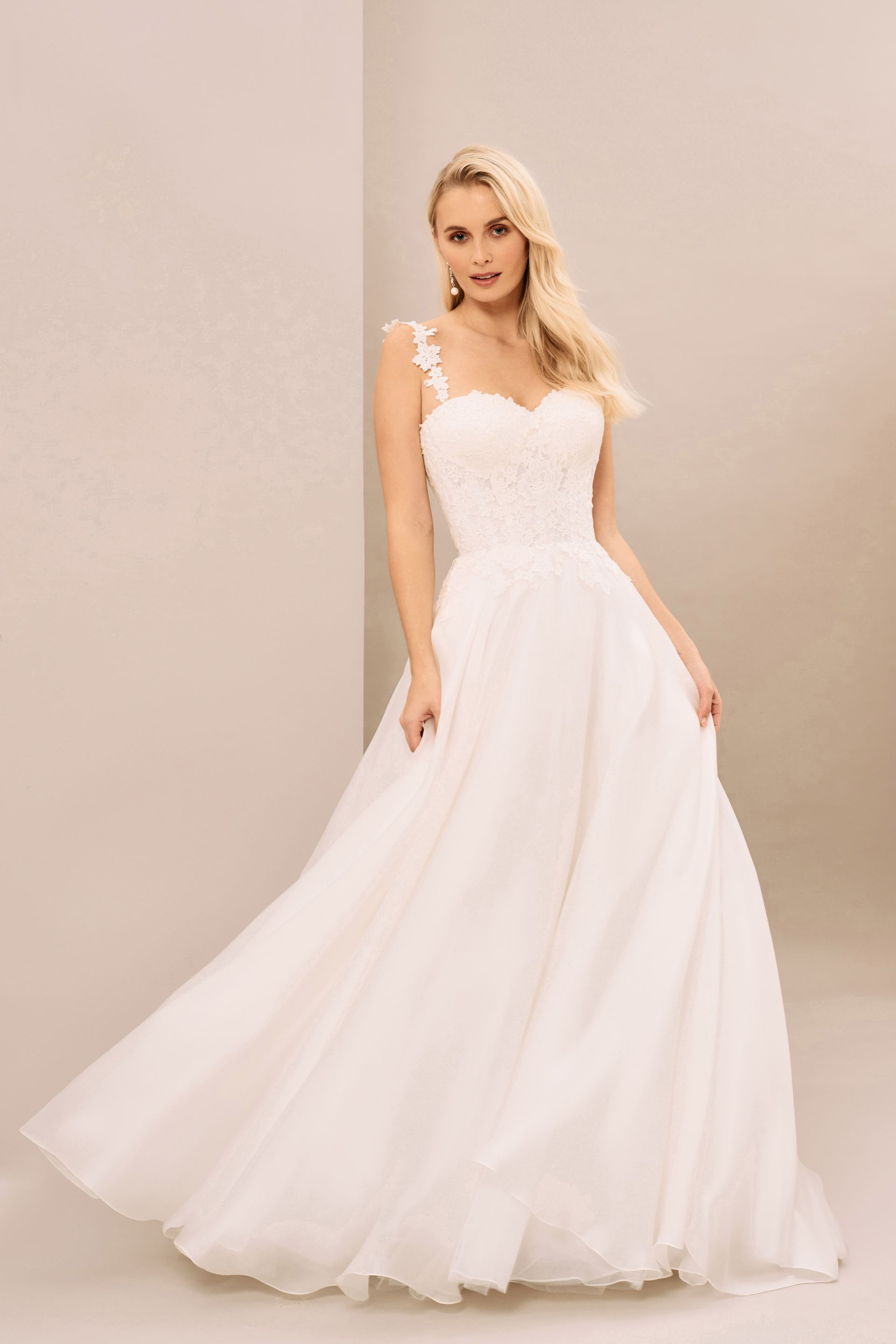 A person wearing a white A-line wedding dress with floral straps and a sweetheart neckline against a neutral backdrop.