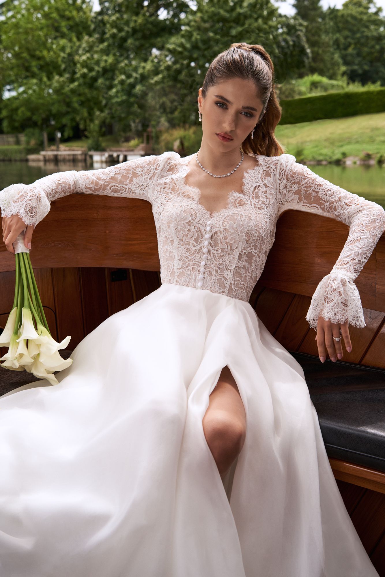 A person in a lace-sleeved wedding gown with a leg slit sits on a wooden boat holding a bouquet of white lilies.