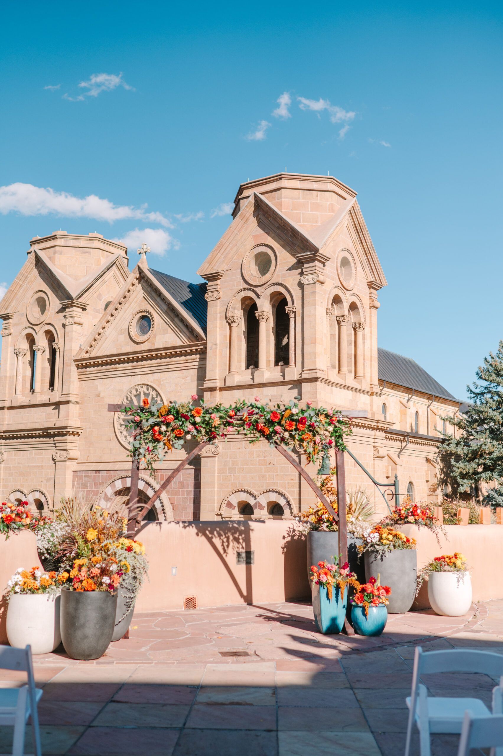 A stone church behind a wedding arch decorated with fall flowers and several large pots of colorful arrangements.