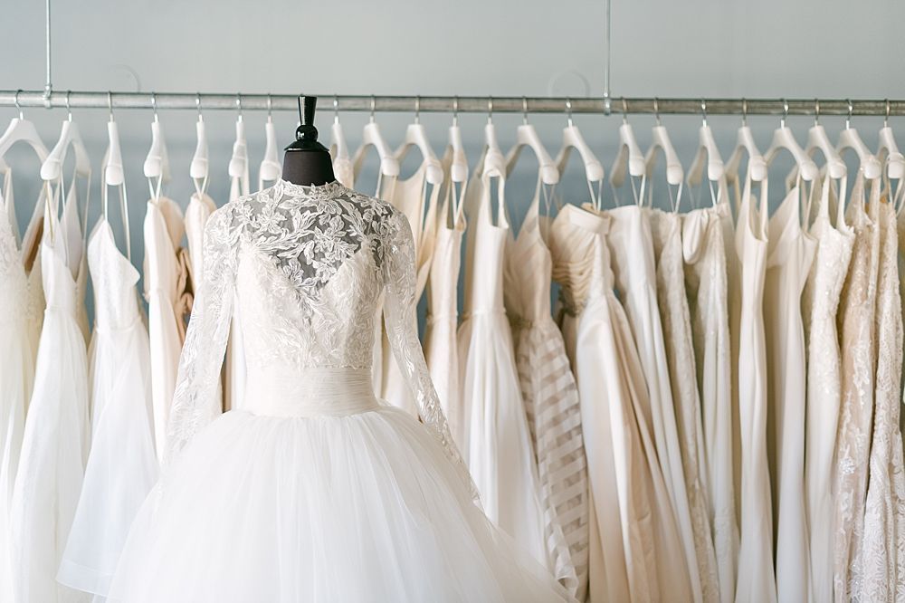 A lace-topped, full-skirted wedding gown on a mannequin, centered in front of a rack of various white bridal dresses.