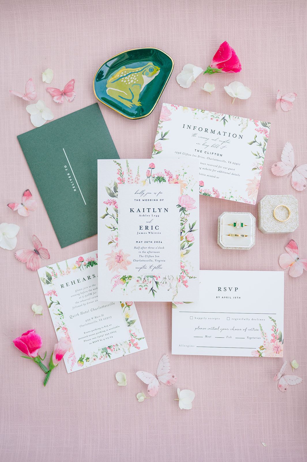 Flat lay of floral-themed wedding stationery on a pink fabric background with an envelope, rings, and decorative accents.