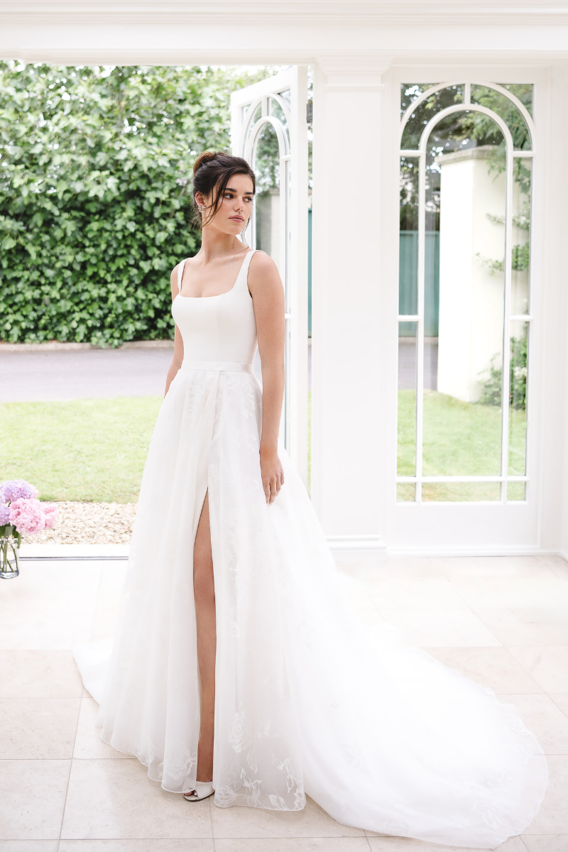 A person wearing a white, sleeveless wedding dress with a square neckline and a high front slit, standing in a sunroom.