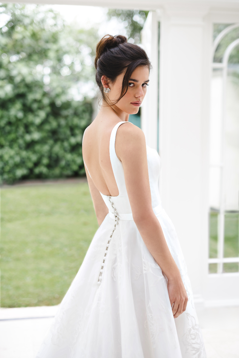 A person in a white backless wedding dress looks over their shoulder while standing in a bright, sunlit room.