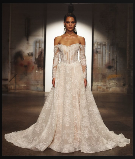A model wears a floor-length, off-the-shoulder lace bridal gown with long sleeves in a dimly lit, industrial studio.