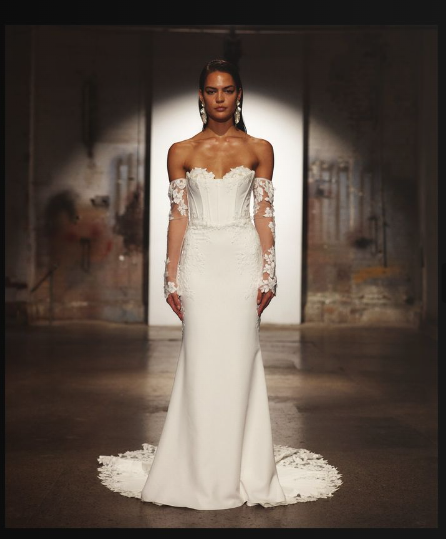 A model in a strapless, floor-length white wedding gown with detachable lace sleeves, posing under a spotlight.