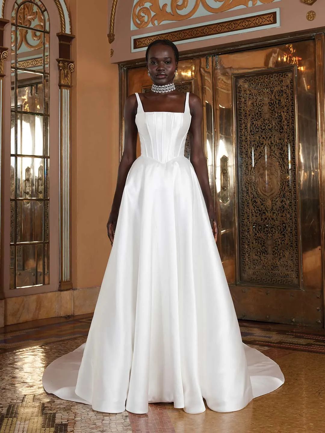 A model wearing a white, floor-length A-line wedding gown with a square neckline and corset bodice in an ornate interior.