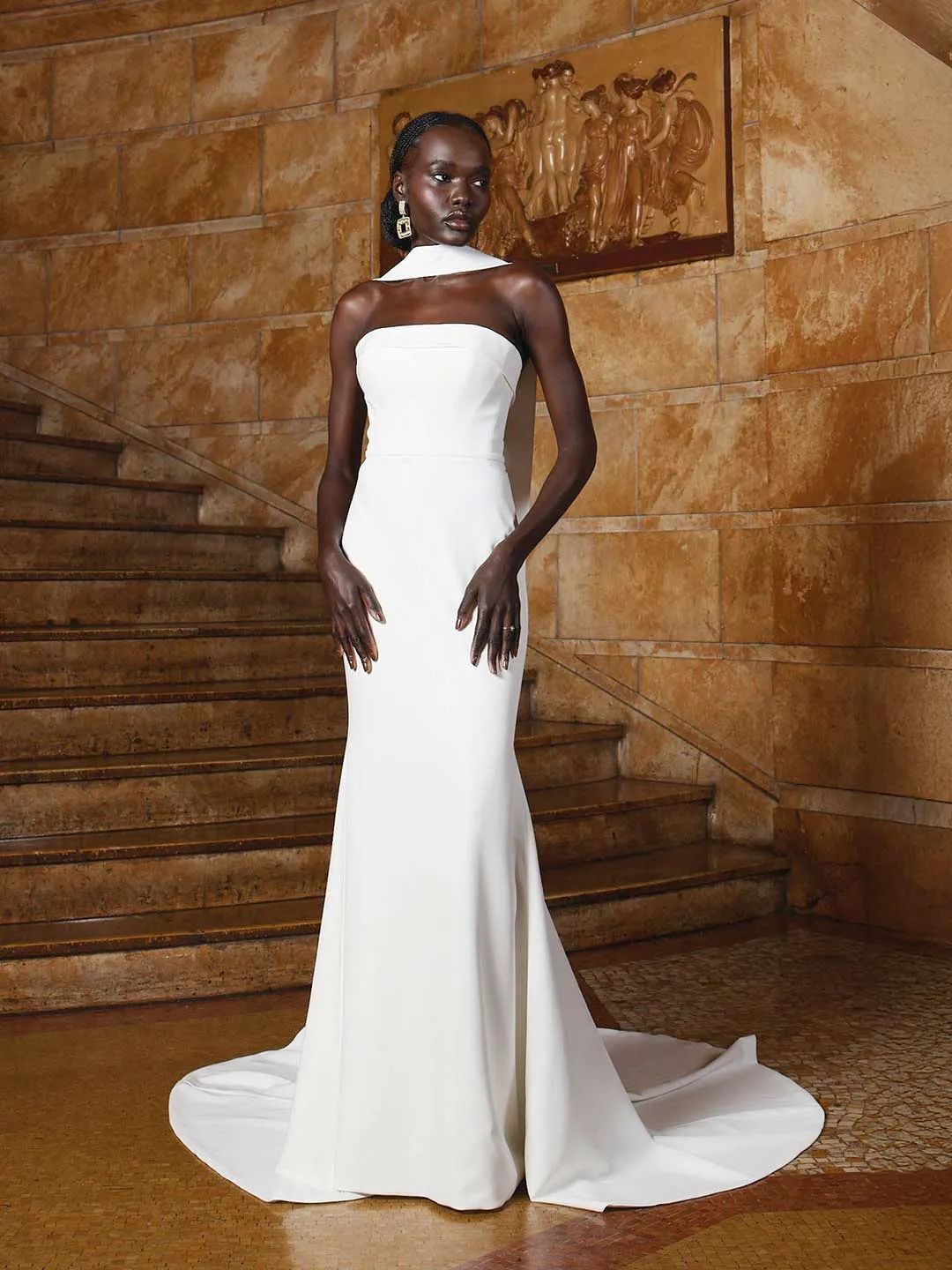 A model wearing a minimalist, strapless white floor-length wedding gown with a train, standing before stone stairs.