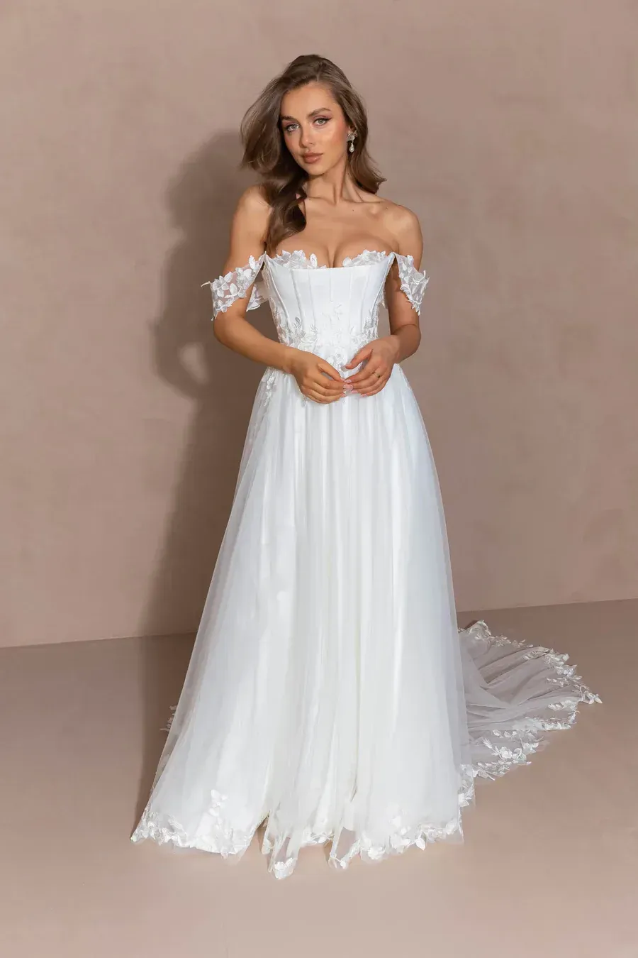 A person wearing a white, off-the-shoulder wedding dress with floral lace detailing, posing against a neutral background.