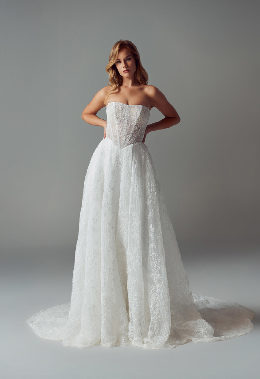 A person wearing a white, strapless, A-line wedding gown with lace detailing, posing against a neutral gray background.