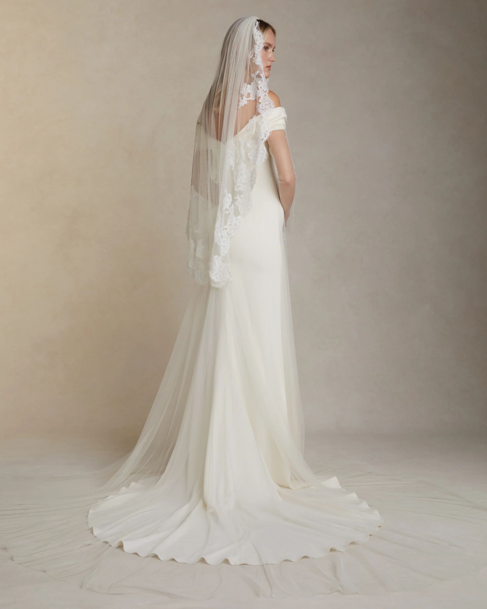 A model stands wearing an off-the-shoulder, floor-length white wedding gown with a train and a lace-trimmed veil.
