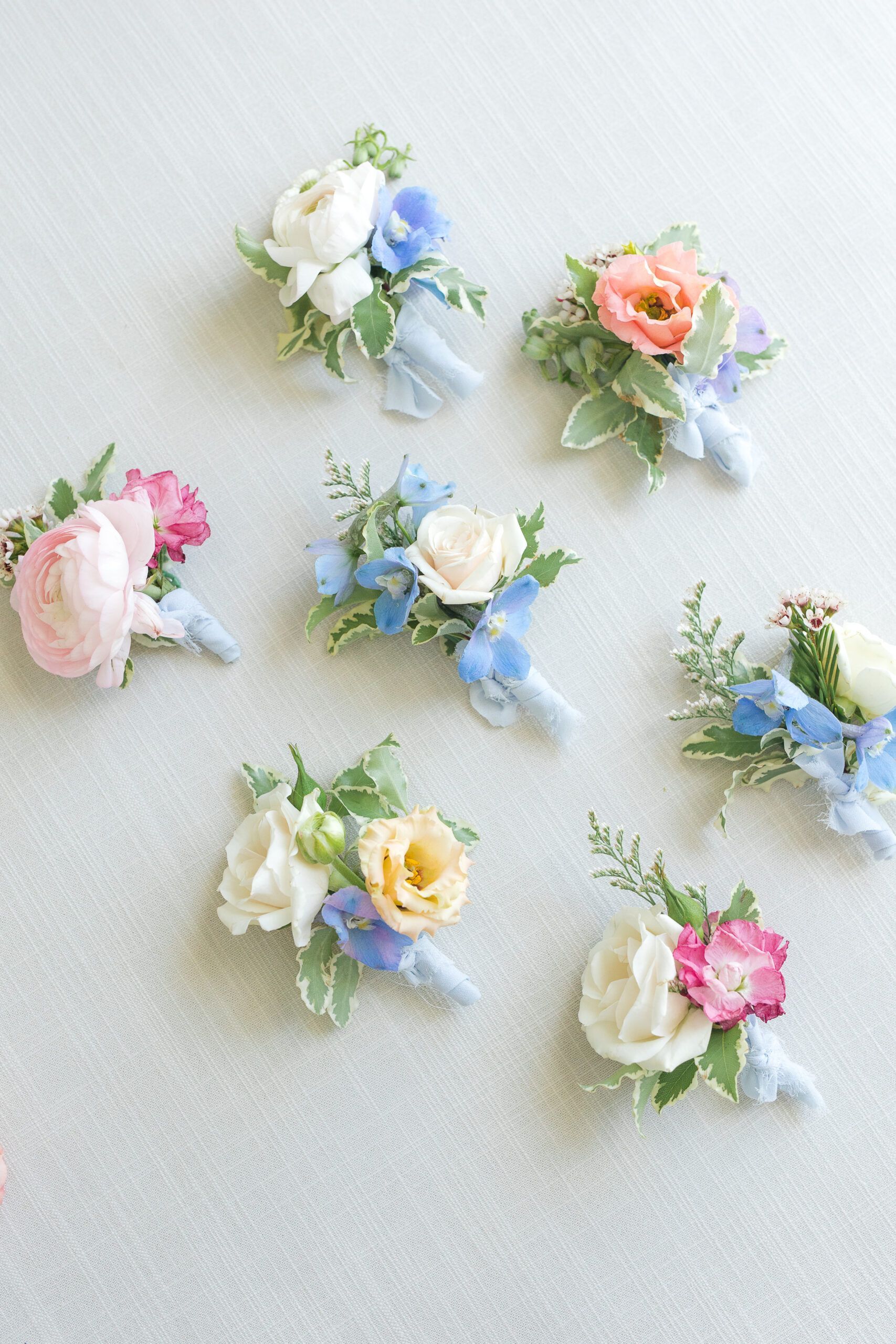 Eight floral boutonnieres featuring roses and blue accent flowers arranged in various pastel color palettes.