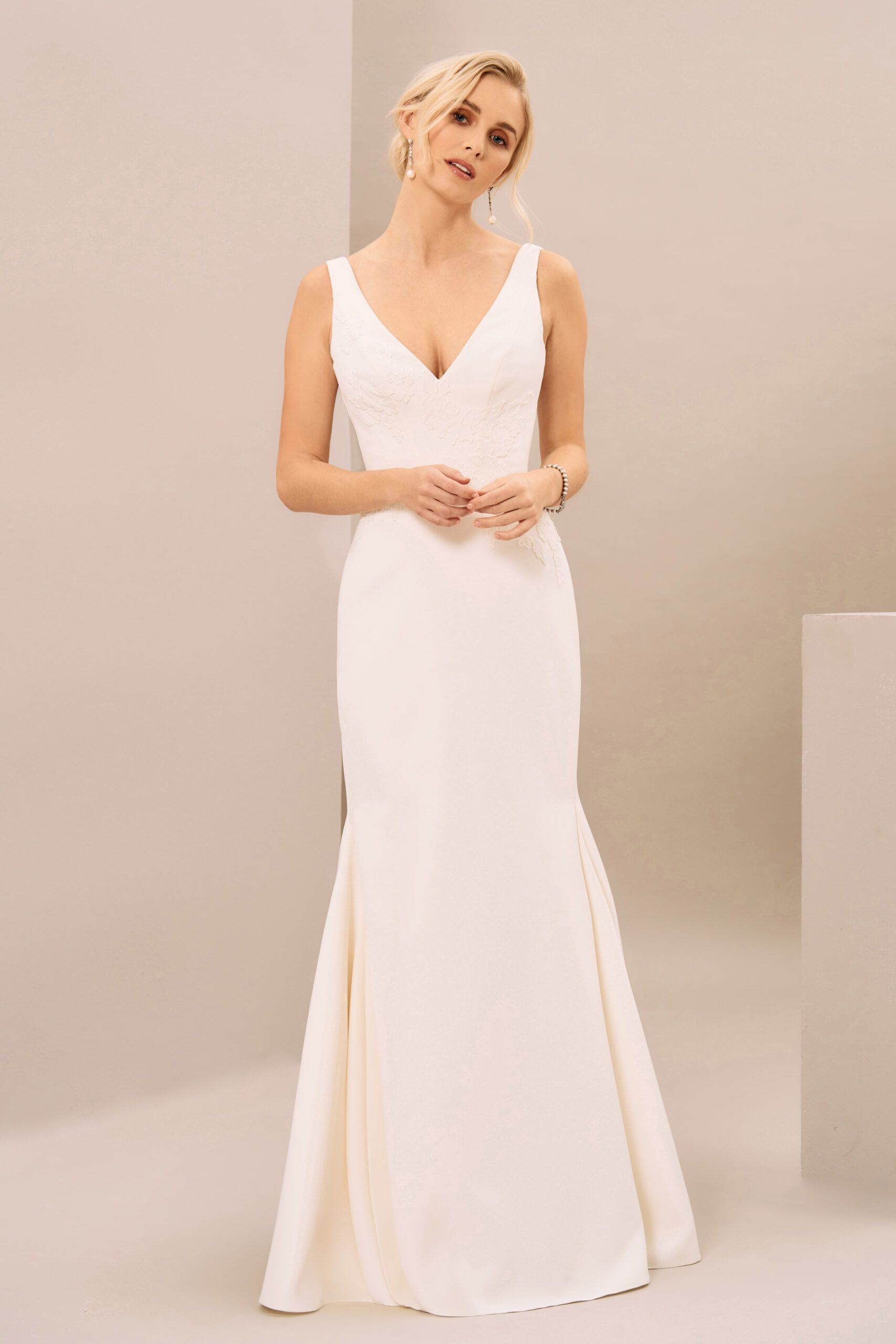 A person in a sleeveless, V-neck white floor-length wedding gown standing against a plain light-colored backdrop.