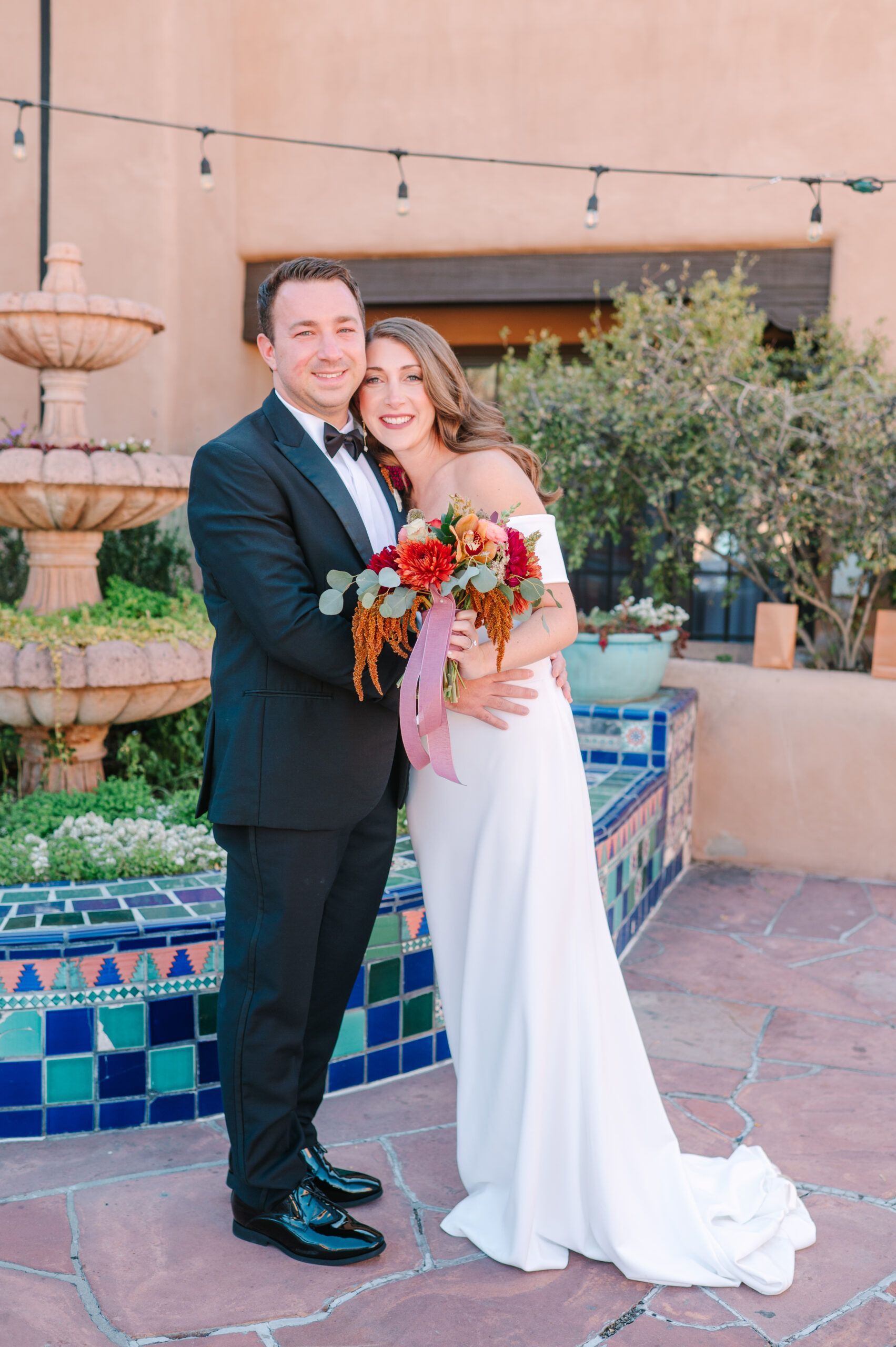 A smiling couple in wedding attire poses together holding a vibrant floral bouquet by an outdoor tiered fountain.