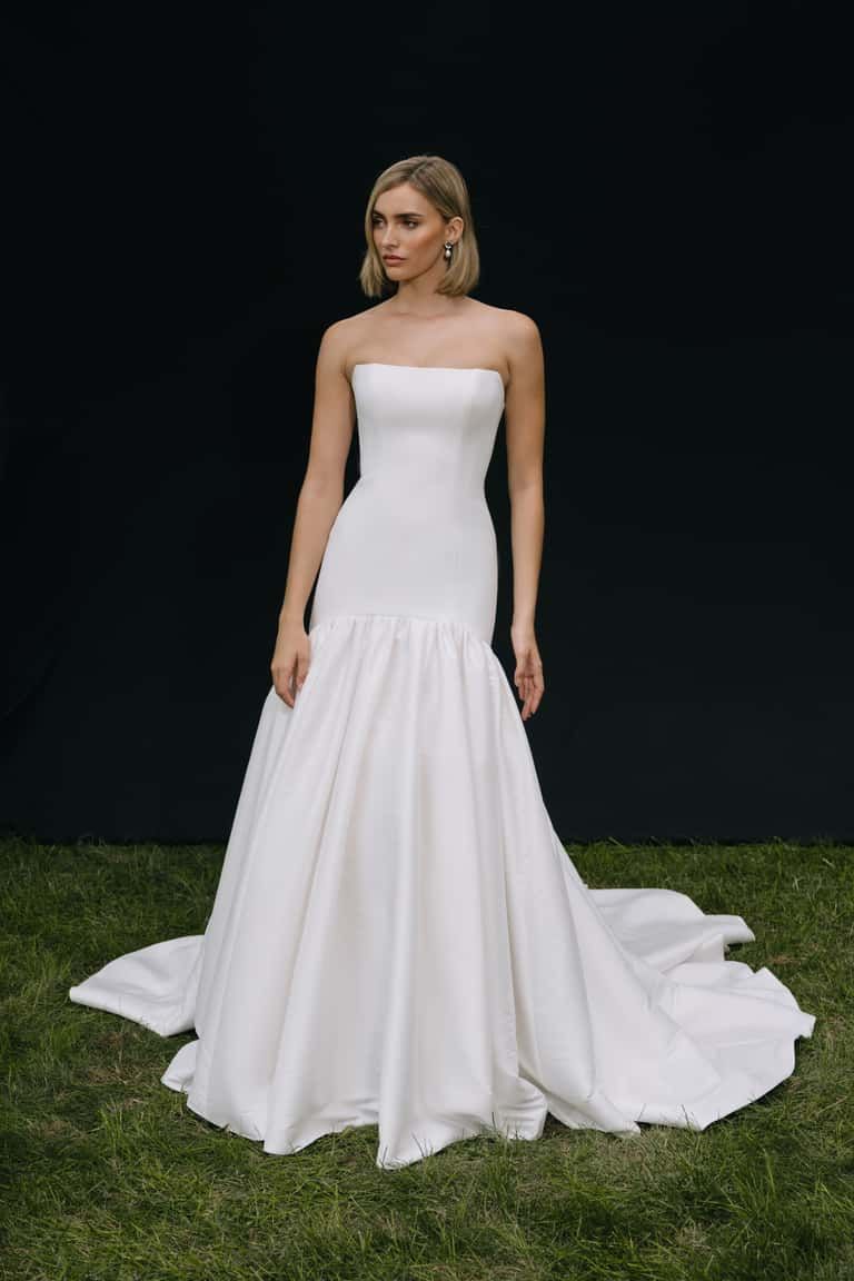 A person wears a strapless, white, floor-length ballgown with a train, standing on grass against a black backdrop.
