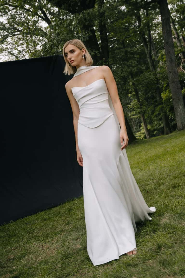 A person in a floor-length white gown with a cowl neckline and attached sheer cape, walking on grass outdoors.
