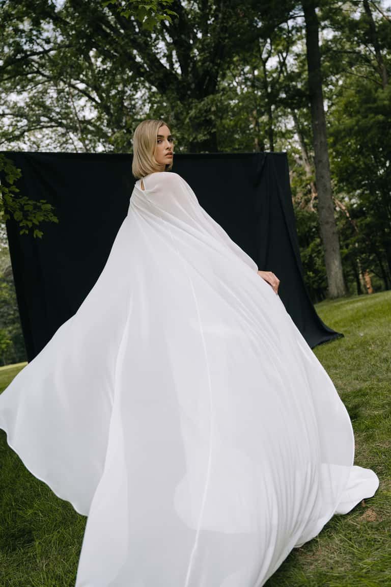A person in a voluminous white cape turns toward the camera against a black backdrop in an outdoor, grassy setting.