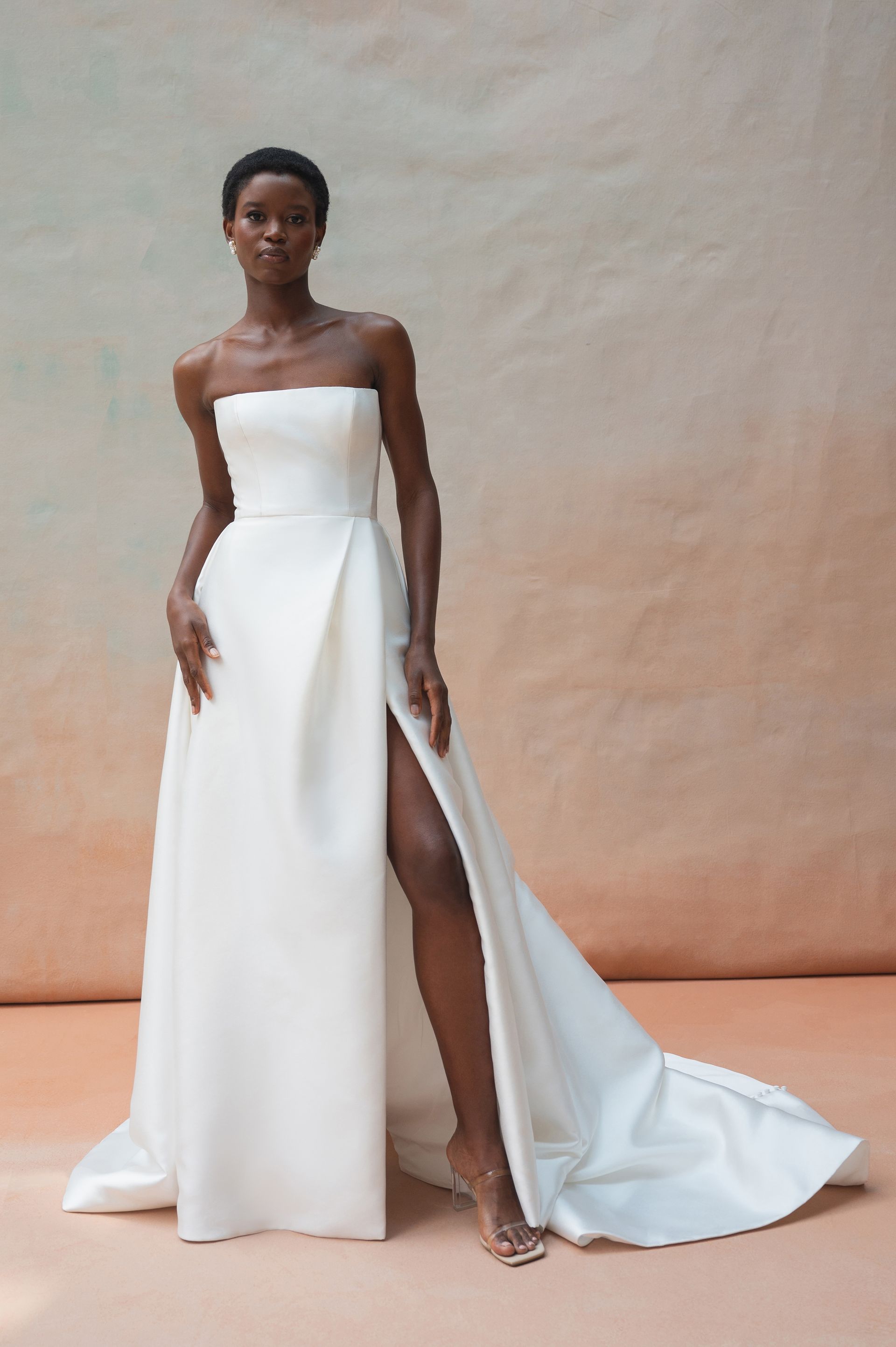 A person models a sleek, strapless white wedding gown with a high leg slit and a train against a beige background.