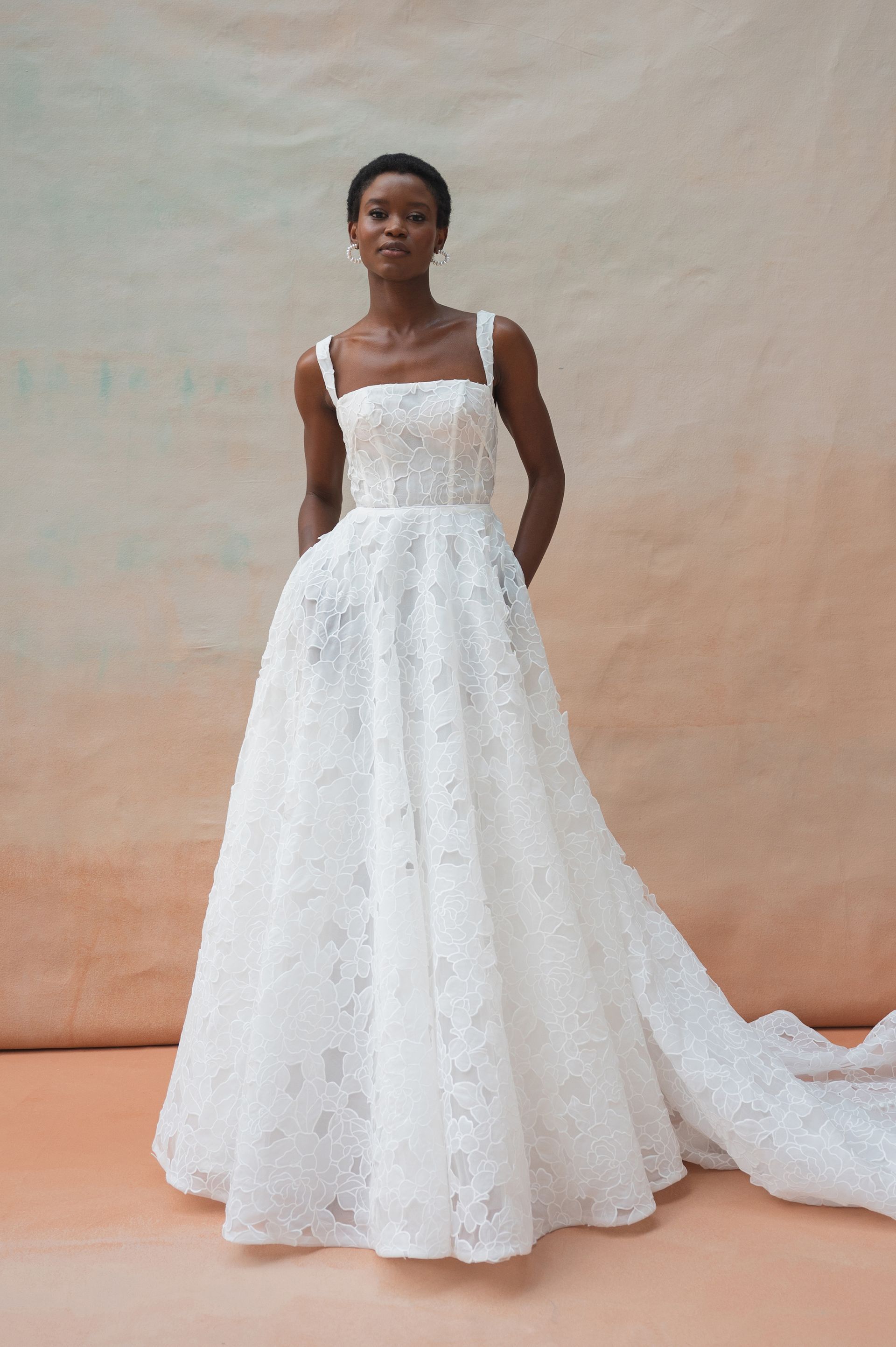 A person wearing a white, floor-length A-line wedding dress with a square neckline and textured floral lace detailing.