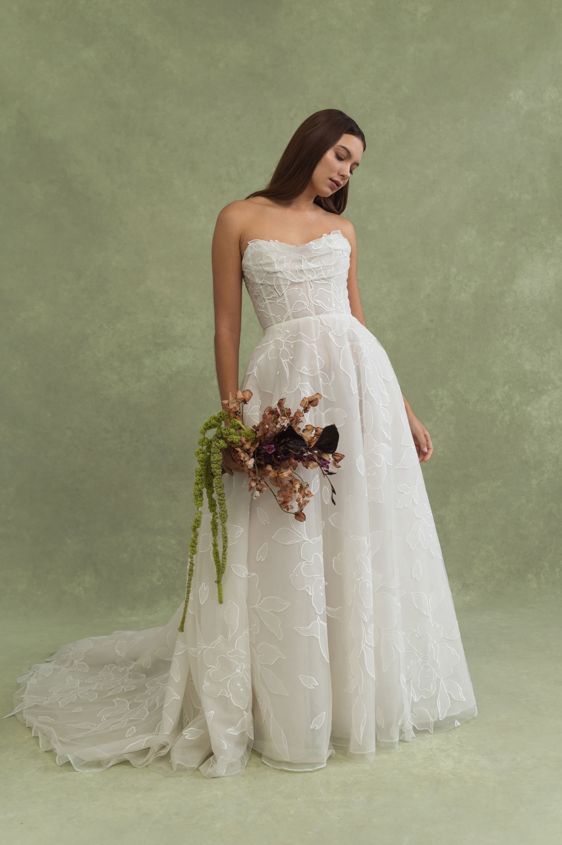 A person wearing a strapless, textured white ballgown wedding dress with a long train, holding a dark floral bouquet.