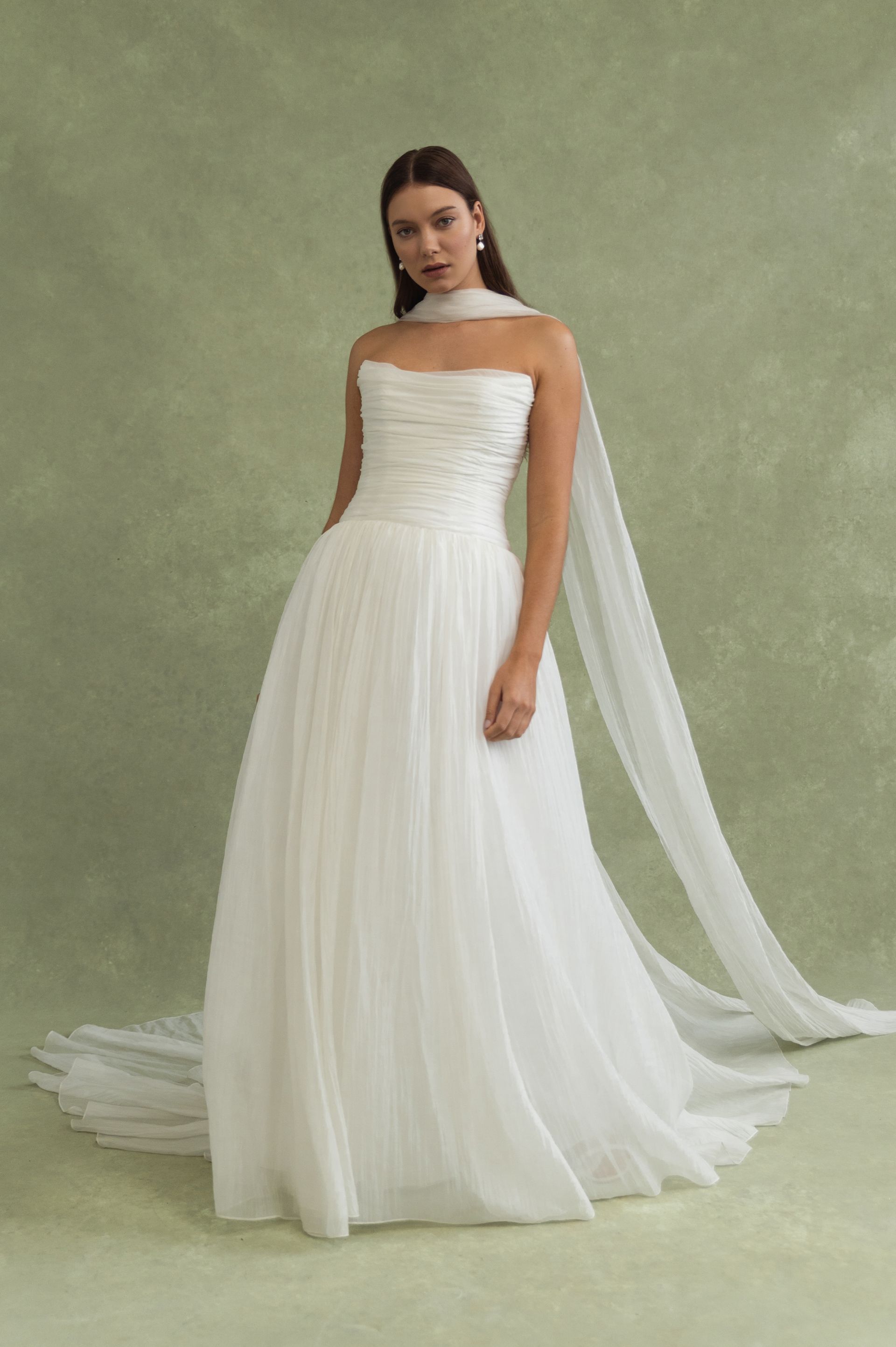 A strapless, floor-length white bridal gown with a structured bodice and a long, flowing sheer scarf draped from the neck.